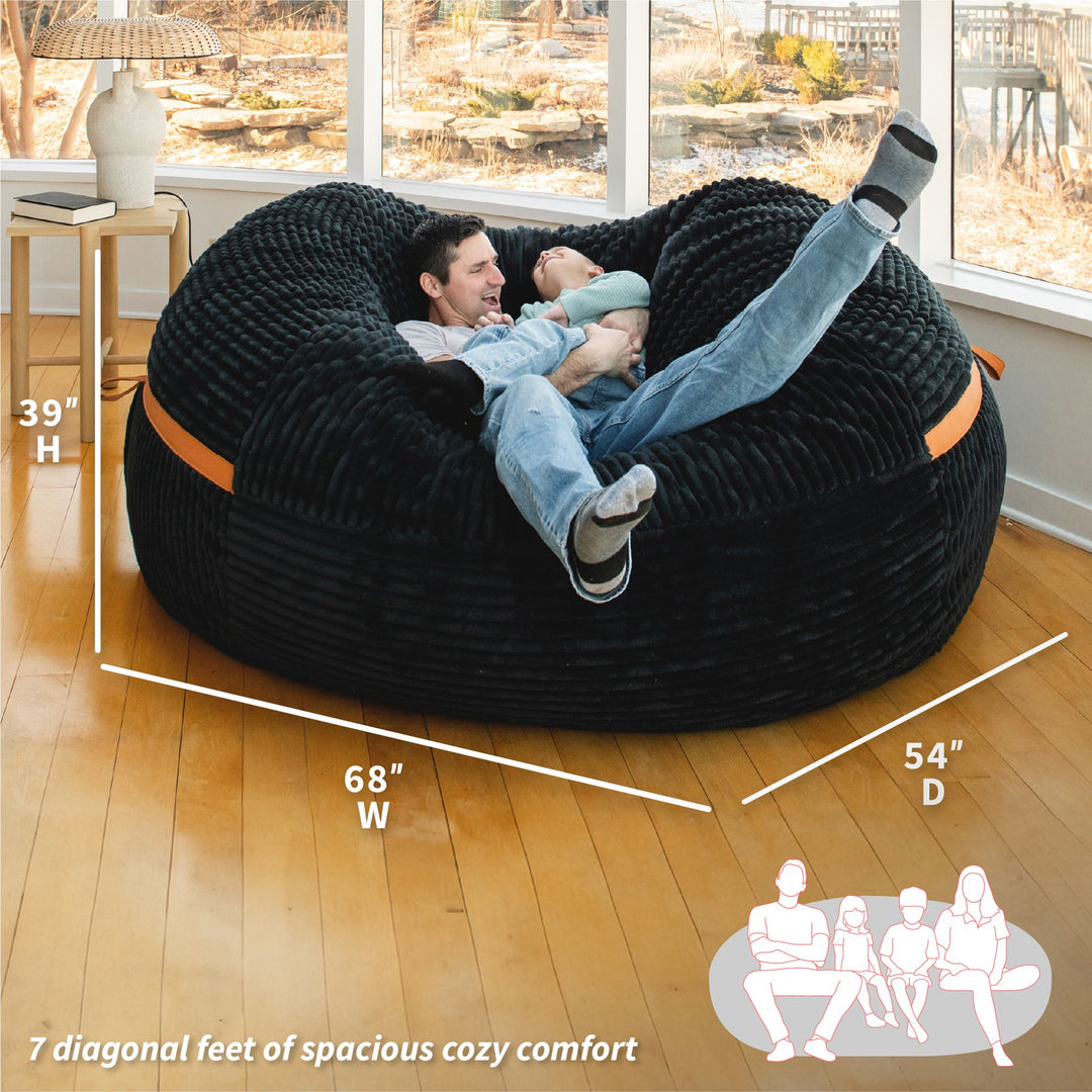 Person lounging in a large black bean bag with measurements and family illustration on a wooden floor. #color_black-licorice-corded-plush