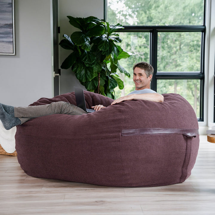 Man using a laptop on a large brown bean bag chair in a room with a plant and window. #color_merlot-cloudweave