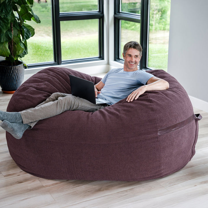 Man sitting on a large brown bean bag chair in a room with large windows. #color_merlot-cloudweave