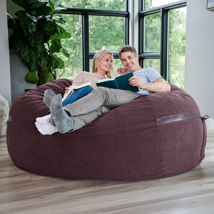 Two people sitting on a large bean bag in a room with large windows and plants. #color_merlot-cloudweave