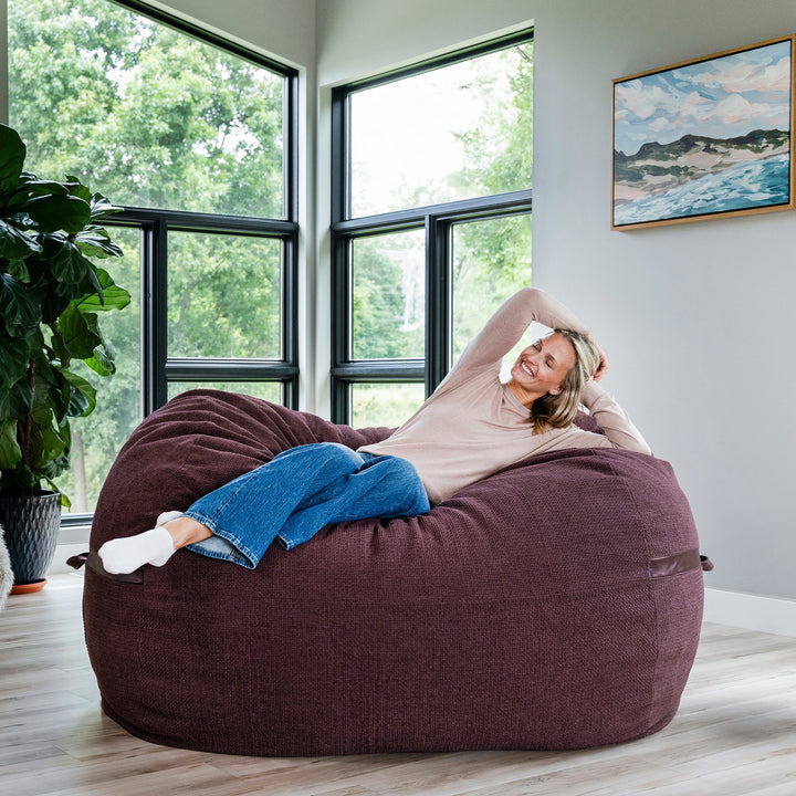 Woman relaxing on a large maroon bean bag chair in a room with large windows and a painting on the wall. #color_merlot-cloudweave