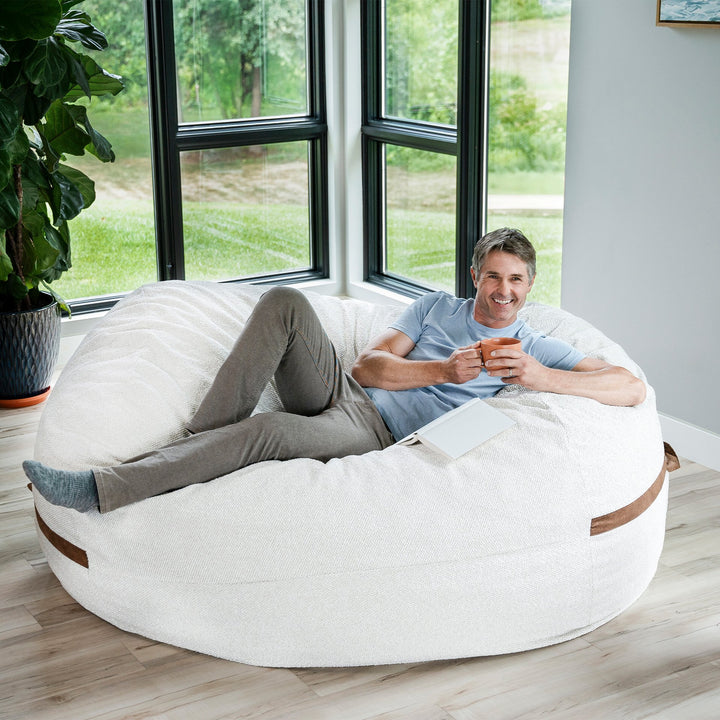 Man relaxing on a large bean bag chair in a bright room with large windows. #color_sandstone-textured-boucle