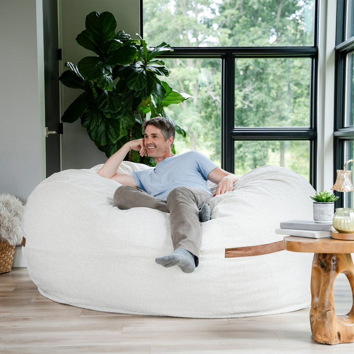 Man relaxing on a white bean bag chair in a bright room with large windows and plants. #color_sandstone-textured-boucle