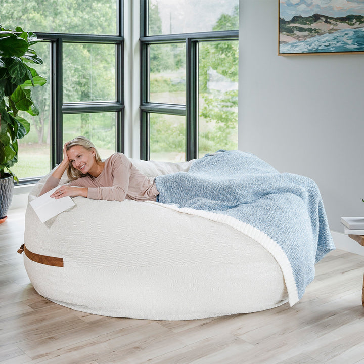 Woman lying on a bean bag chair with a blue blanket in a bright room with large windows. #color_sandstone-textured-boucle