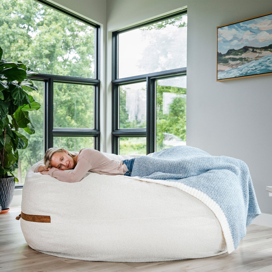 Person lying on a bean bag chair with a blanket in a room with large windows and a plant. #color_sandstone-textured-boucle