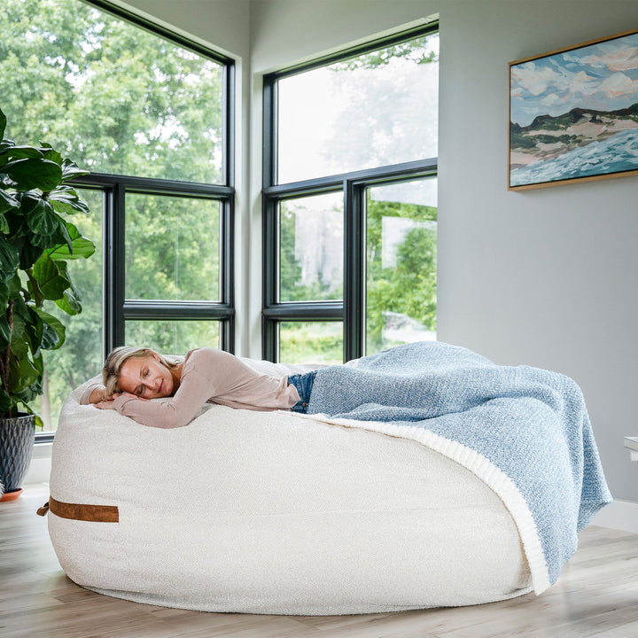 Person lying on a bean bag chair with a blanket in a room with large windows and a plant. #color_sandstone-textured-boucle