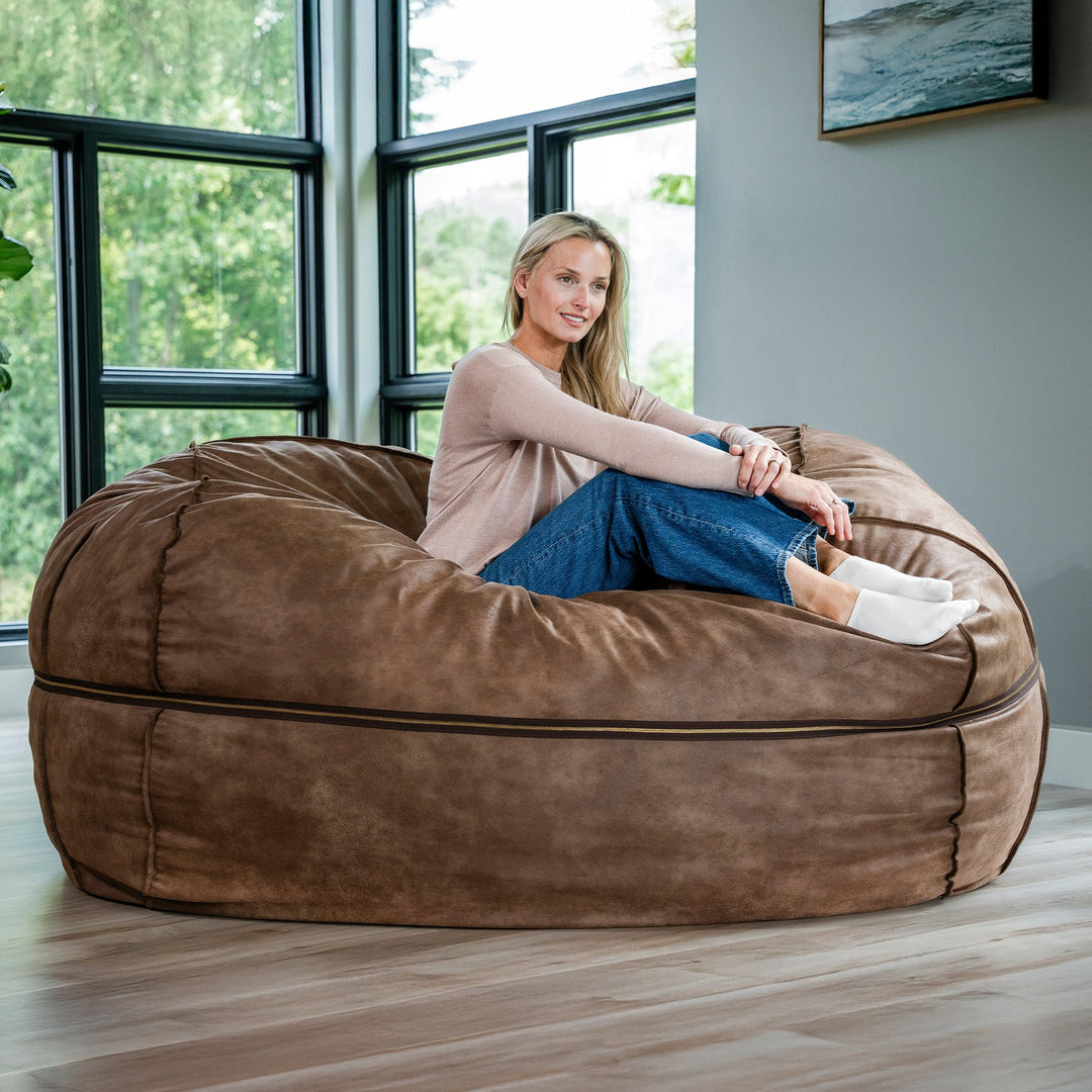 Woman sitting on a large brown bean bag chair in a modern room with large windows. #color_coffee-soft-vegan-leather