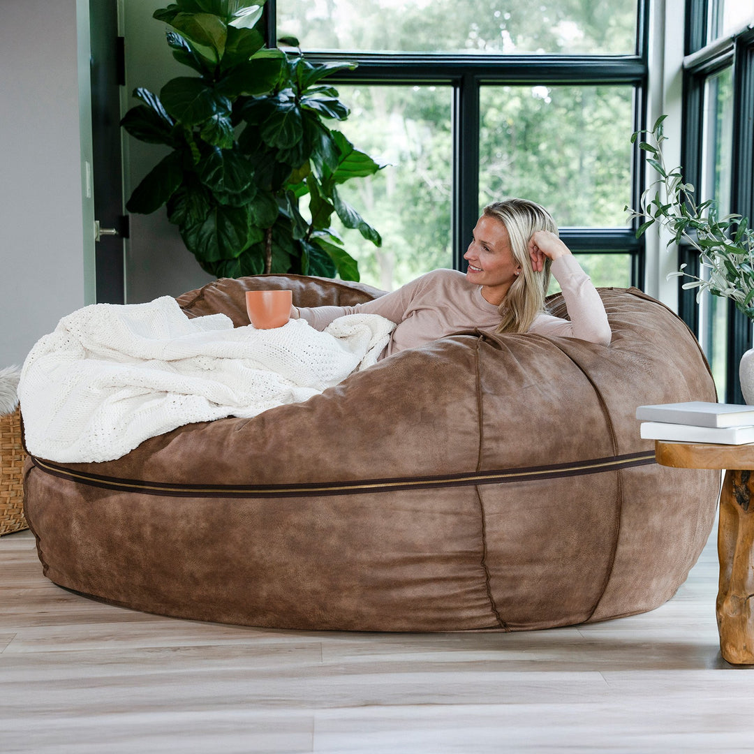 Woman relaxing on a large brown bean bag chair in a bright room with plants. #color_coffee-soft-vegan-leather