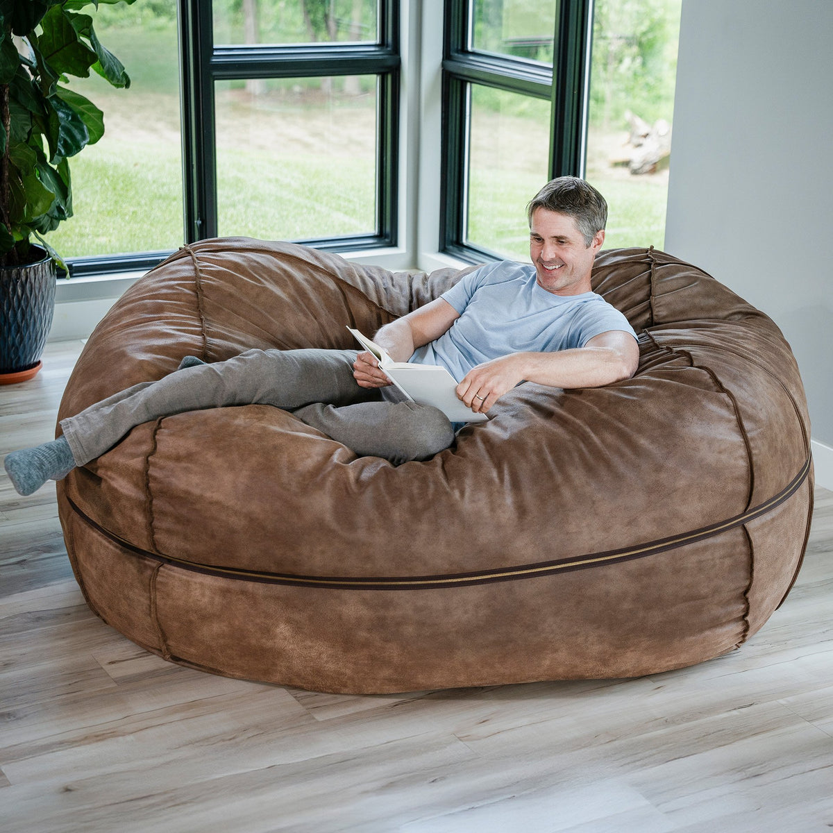 Man sitting on a large brown bean bag chair reading a book in a room with large windows. 