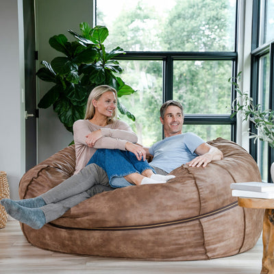 Two people sitting on a large brown bean bag chair in a room with large windows and plants. 