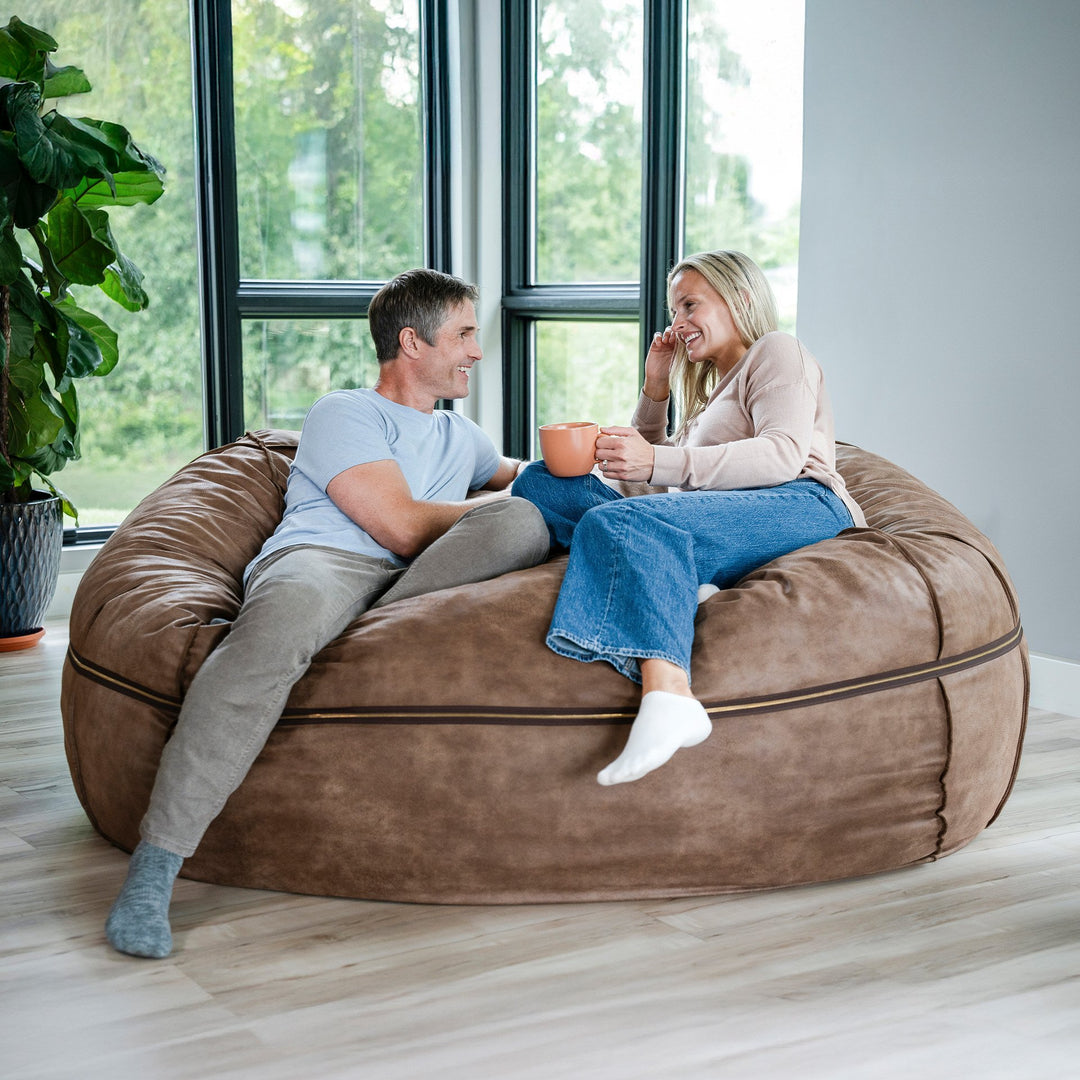 Two people sitting on a large brown bean bag in a room with large windows. #color_coffee-soft-vegan-leather