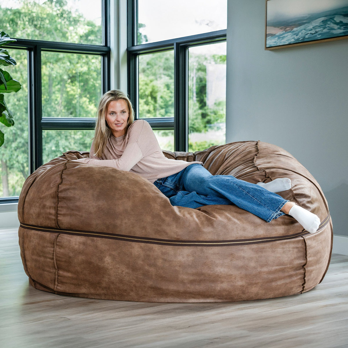Woman sitting on a large brown bean bag chair in a room with large windows. 