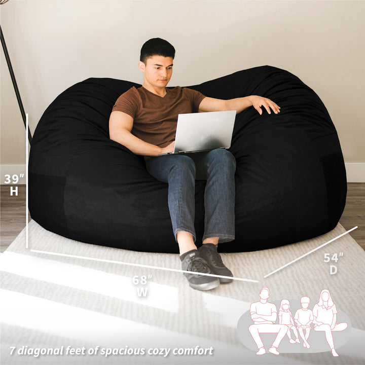 Person using a laptop on a large black bean bag chair with measurements displayed. #color_black-plush