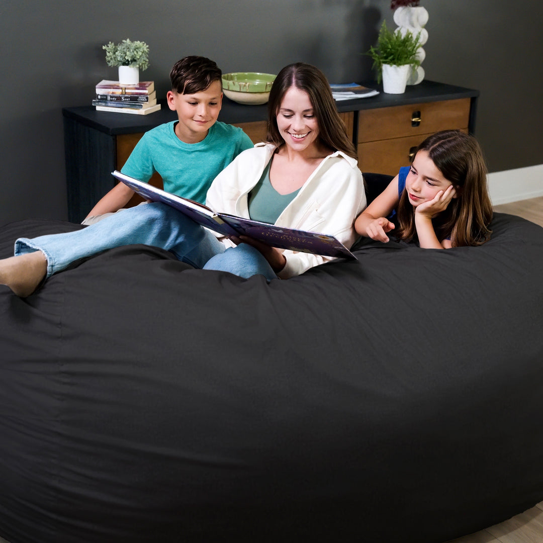 Woman and two children sitting on a large black bean bag reading a book together. #color_black-brushed-denim