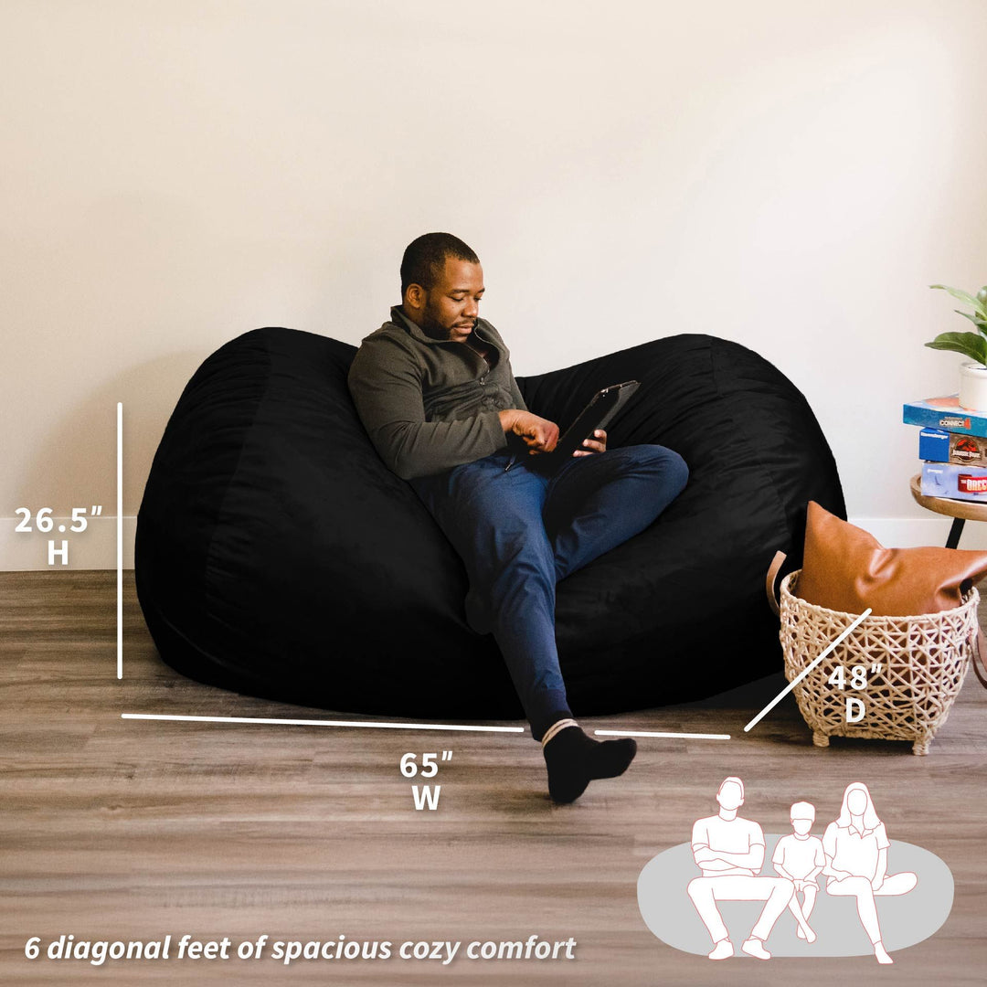 Man sitting on a large black bean bag chair with measurements and comfort information displayed. #color_black-plush