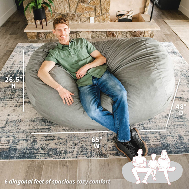 Man sitting on a large gray bean bag chair with dimensions displayed, in a living room setting. #color_fog-brushed-denim
