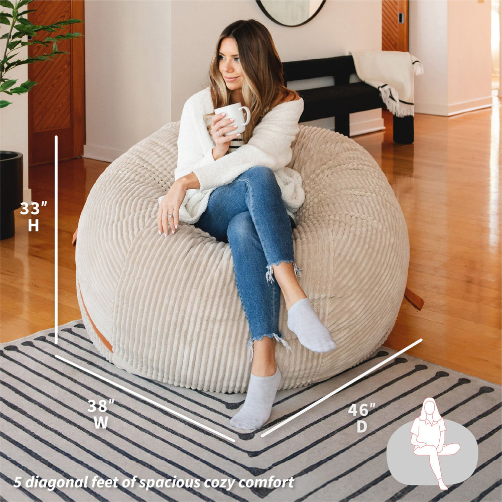 Woman sitting on a bean bag chair holding a mug, with measurements displayed. #color_vanilla-latte-corded-plush