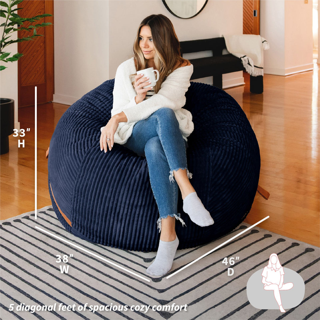 Woman sitting on a navy bean bag chair holding a mug, with measurements displayed. #color_midnight-blue-corded-plush