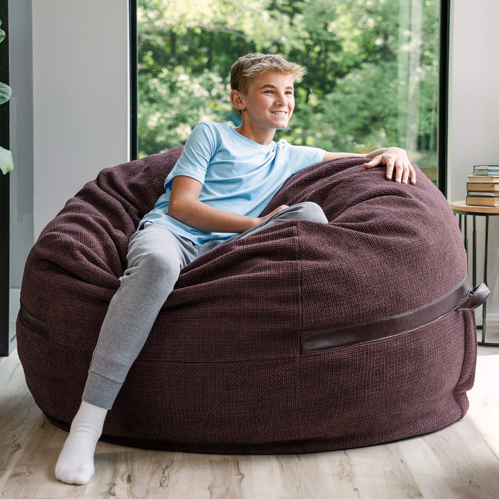 Young boy sitting on a large maroon bean bag chair indoors with a view of greenery outside. #color_merlot-cloudweave