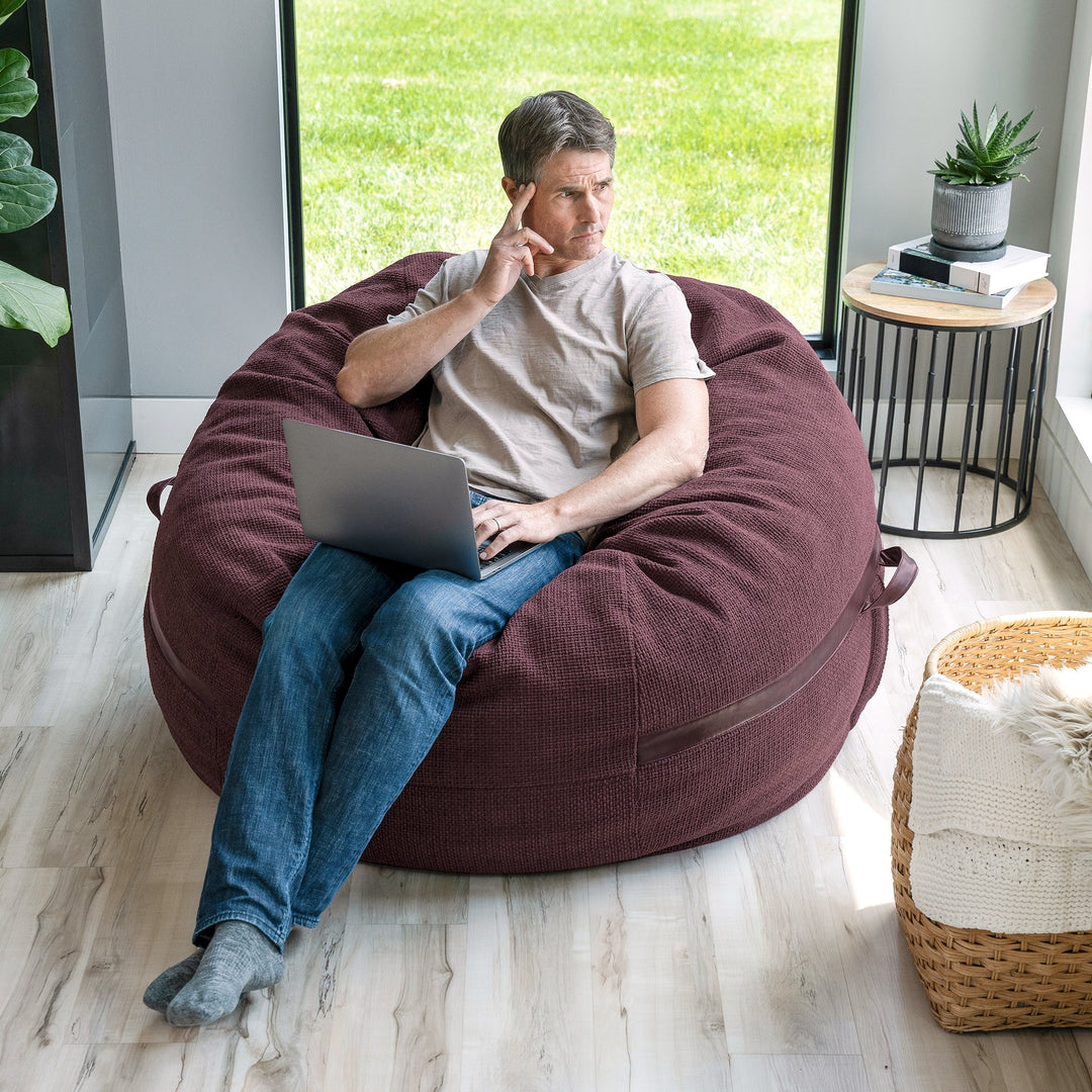 Man sitting on a large burgundy bean bag chair using a laptop in a modern room. #color_merlot-cloudweave