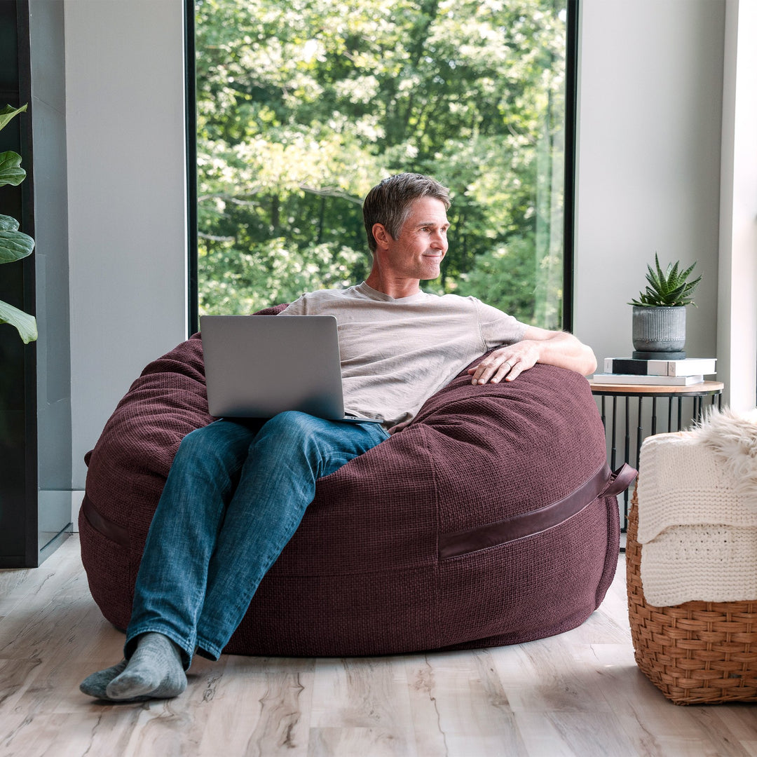Man sitting on a bean bag chair using a laptop in a modern living room. #color_merlot-cloudweave