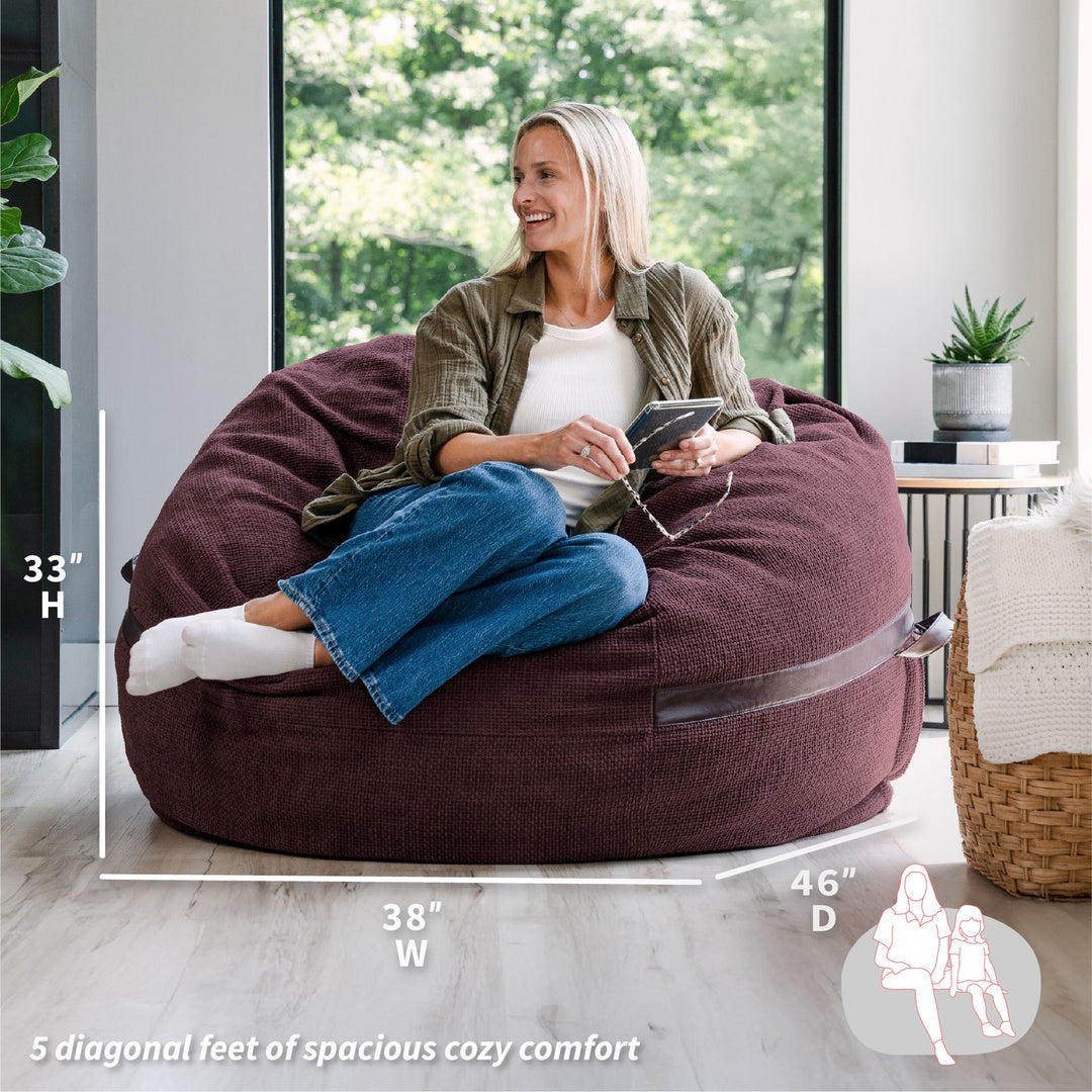 Woman sitting on a large brown bean bag chair in a modern living room with dimensions displayed. #color_merlot-cloudweave