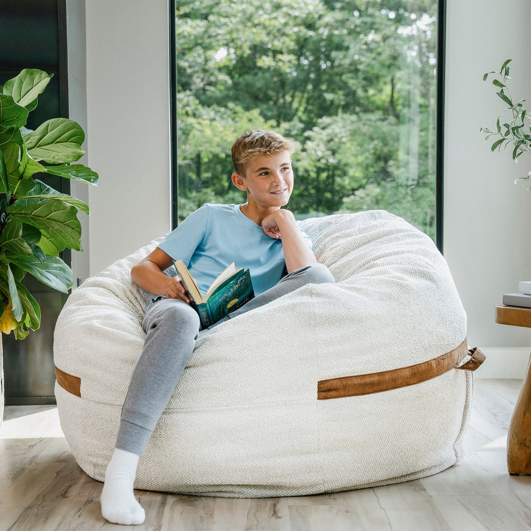 Young boy sitting on a bean bag chair reading a book in a room with large windows and plants. #color_sandstone-textured-boucle