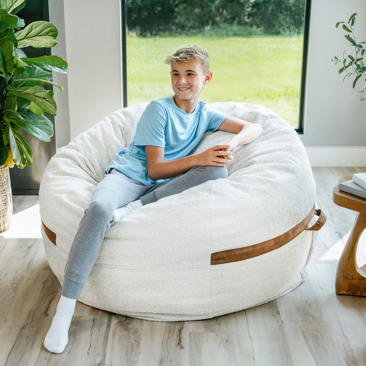 Young boy sitting on a bean bag chair in a bright room with plants and a window. #color_sandstone-textured-boucle