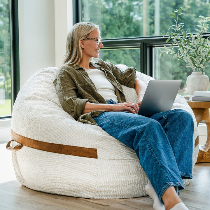 Woman using a laptop on a large bean bag chair in a bright room with large windows. #color_sandstone-textured-boucle