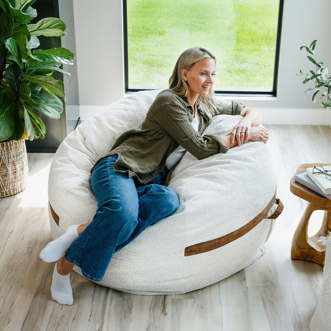 Woman sitting on a bean bag chair in a bright room with plants and a window. #color_sandstone-textured-boucle