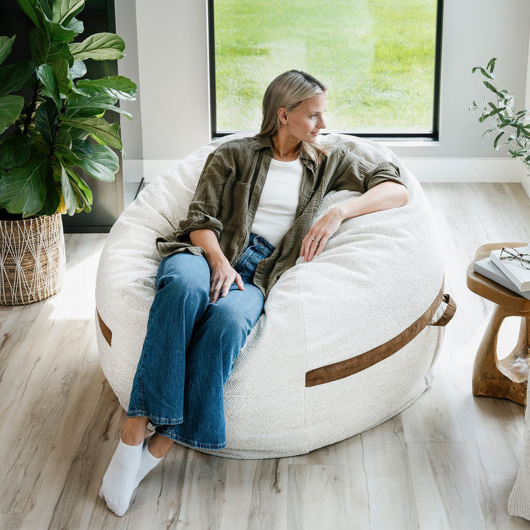 Woman sitting on a bean bag chair in a bright room with plants and a window. #color_sandstone-textured-boucle