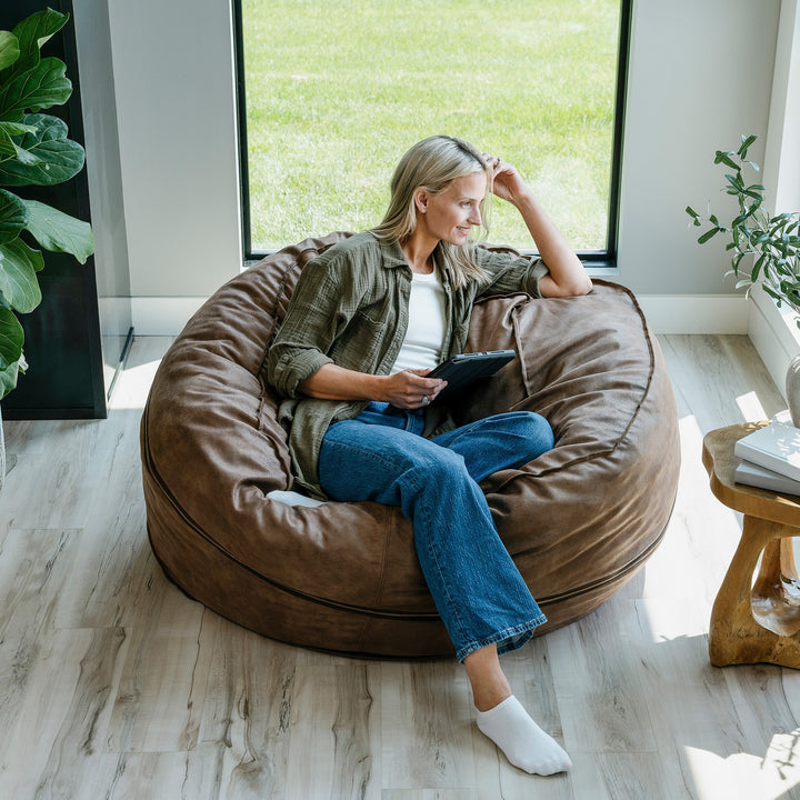 Woman sitting on a brown bean bag chair indoors, holding a tablet. #color_coffee-soft-vegan-leather