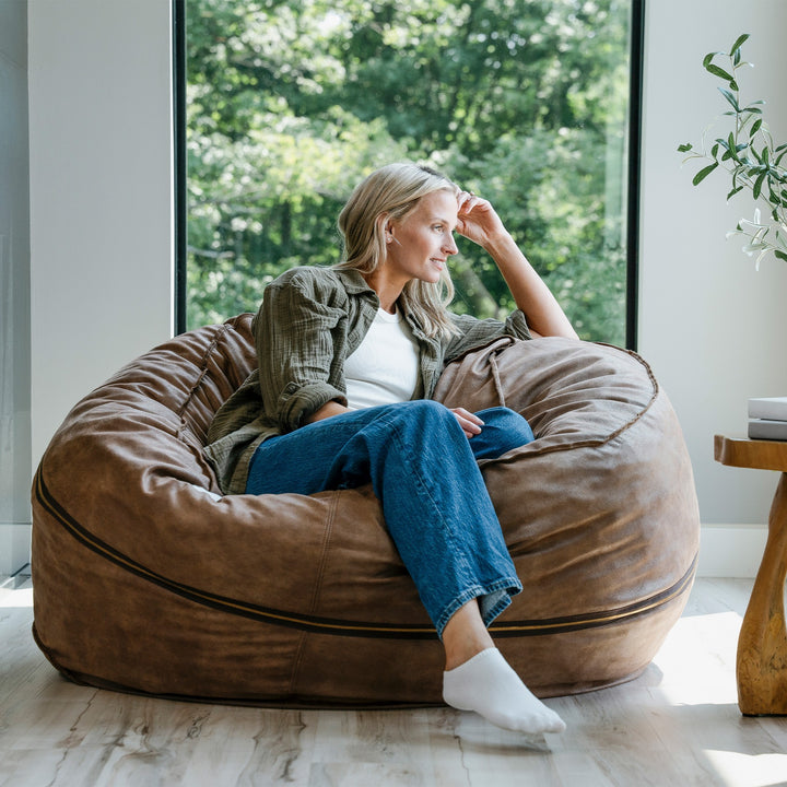 Woman sitting on a large brown bean bag chair in a room with large windows and greenery outside. #color_coffee-soft-vegan-leather