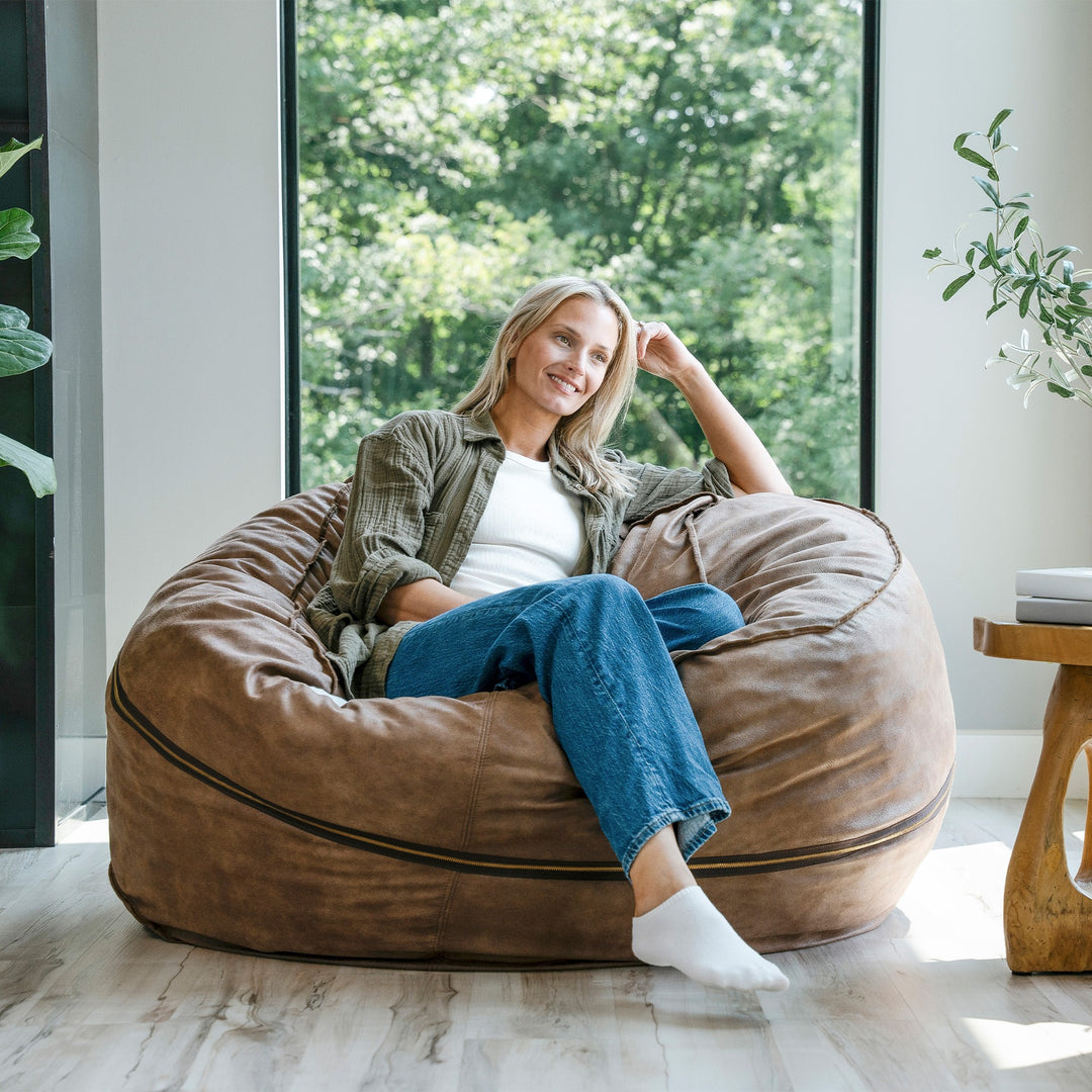 Woman sitting on a large brown bean bag chair in a bright room with large windows. #color_coffee-soft-vegan-leather