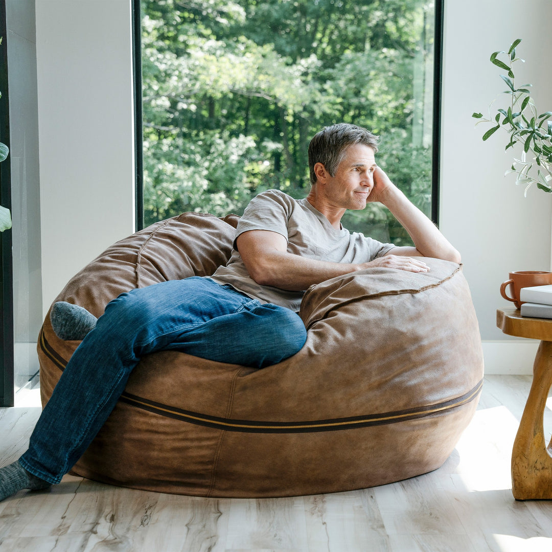 Man sitting on a large brown bean bag chair in a bright room with large windows. #color_coffee-soft-vegan-leather