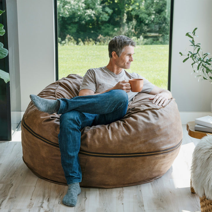 Man sitting on a brown bean bag chair holding a mug, with a view of greenery outside. #color_coffee-soft-vegan-leather