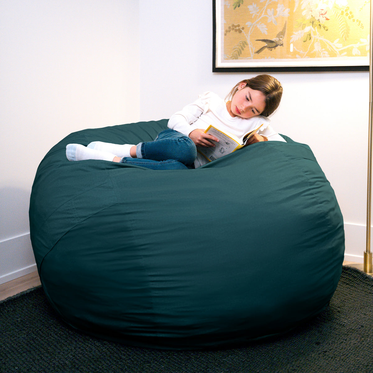 Child lying on a large green bean bag reading a book in a room with a painting on the wall. 