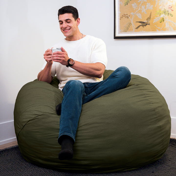 Man sitting on a large green bean bag chair using a smartphone in a room with a painting on the wall. #color_forest-green-velveteen