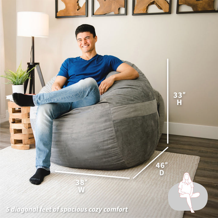 Man sitting on a gray bean bag chair with dimensions displayed, in a room setting. #color_gray-plush