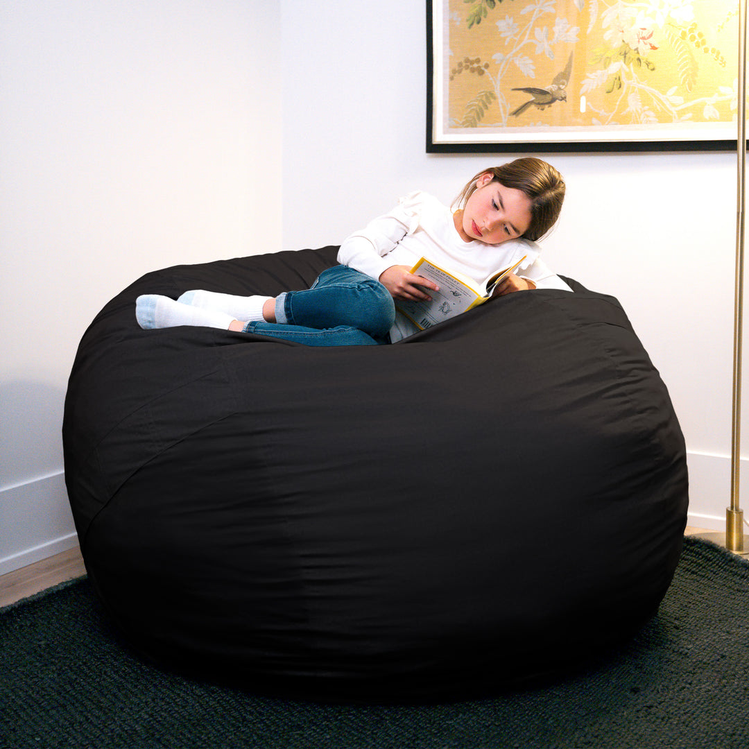 Person lying on a large black bean bag reading a book in a room with a floral painting on the wall. #color_black-brushed-denim