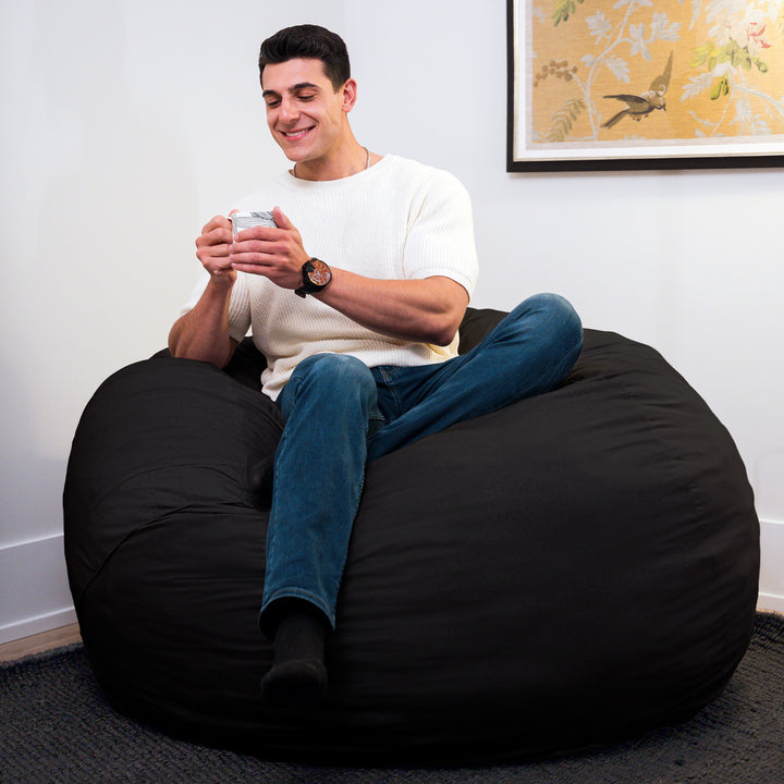 Man sitting on a black bean bag chair holding a phone in a room with a floral painting on the wall. #color_black-brushed-denim