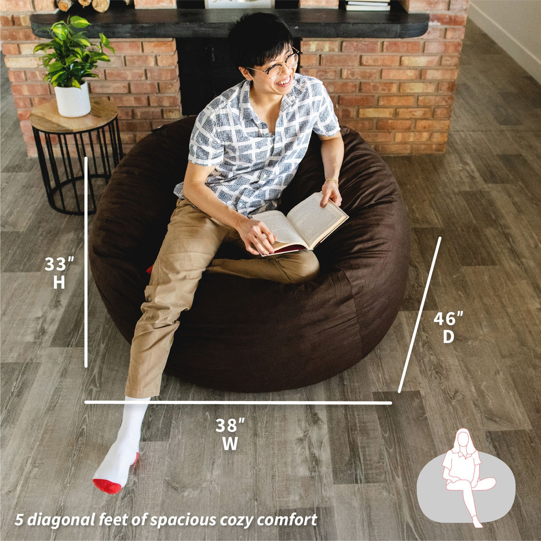 Man sitting on a bean bag chair reading a book with measurements displayed. #color_cocoa-brushed-denim