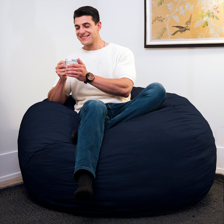 Man sitting on a navy bean bag chair holding a phone, with a white wall and framed picture in the background. #color_cobalt-brushed-denim