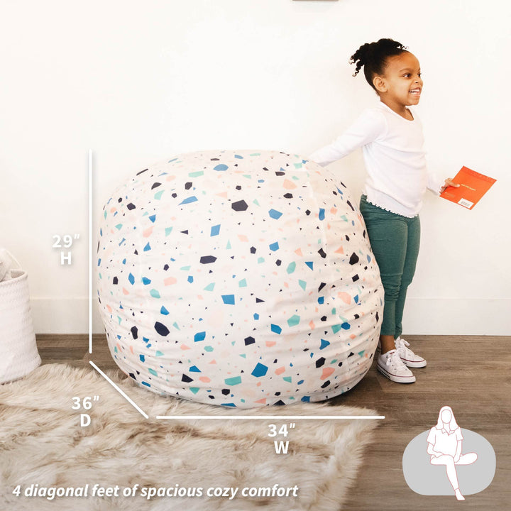 Child standing next to a large bean bag with measurements and text on a wooden floor. #color_dolce-terrazzo-brushed-denim