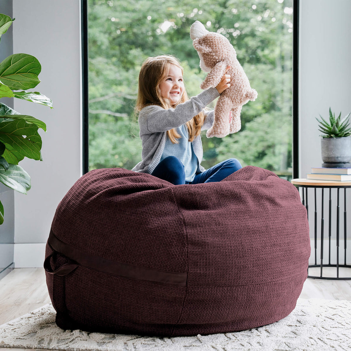 Child playing with a teddy bear on a merlot brown bean bag chair in a room with a window and plants. 