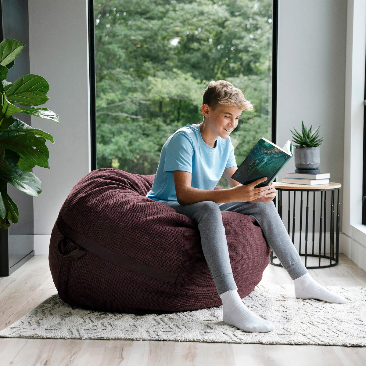 Child sitting on a bean bag reading a book in a bright room with large windows. 