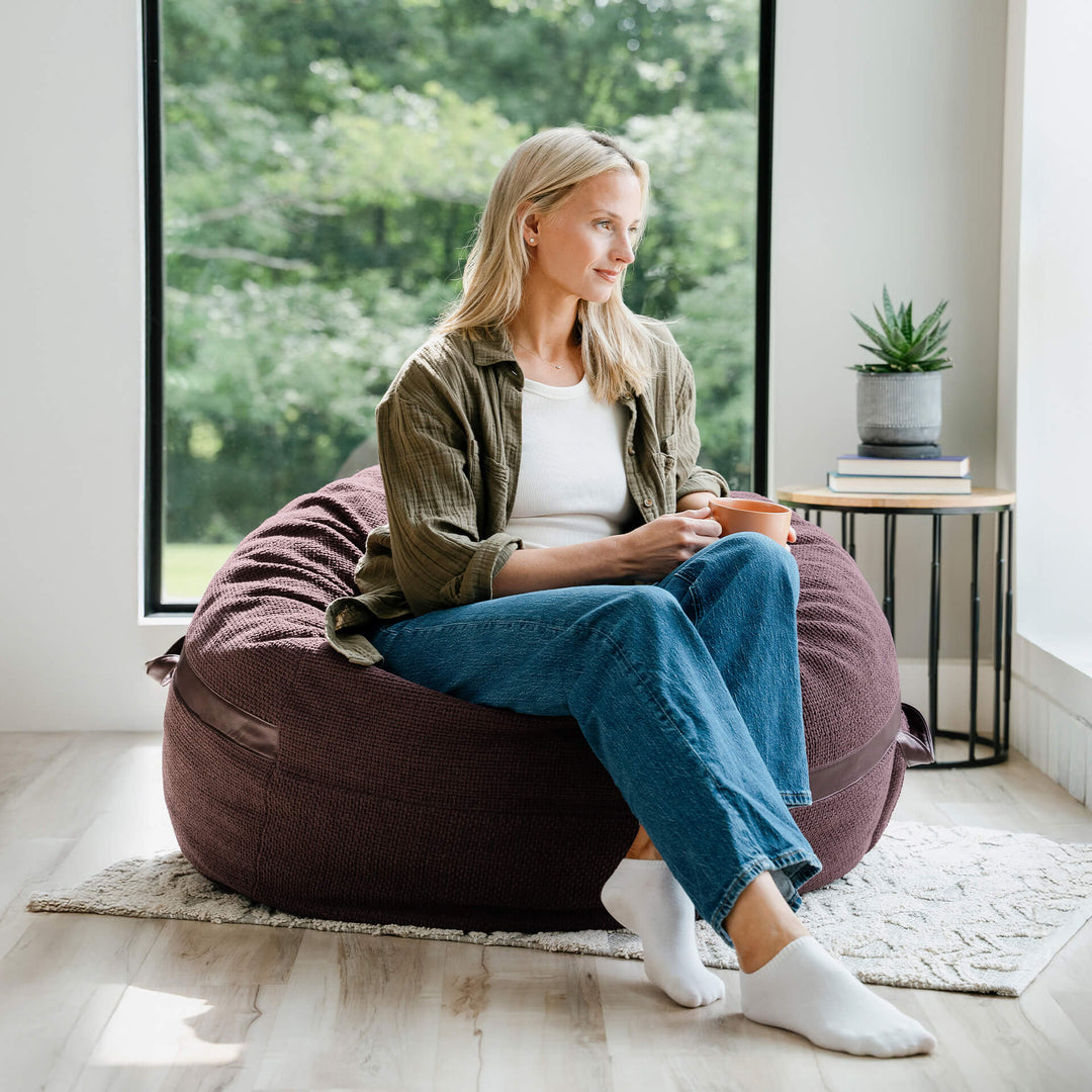 Woman sitting on a bean bag chair in a bright room with large windows. #color_merlot-cloudweave