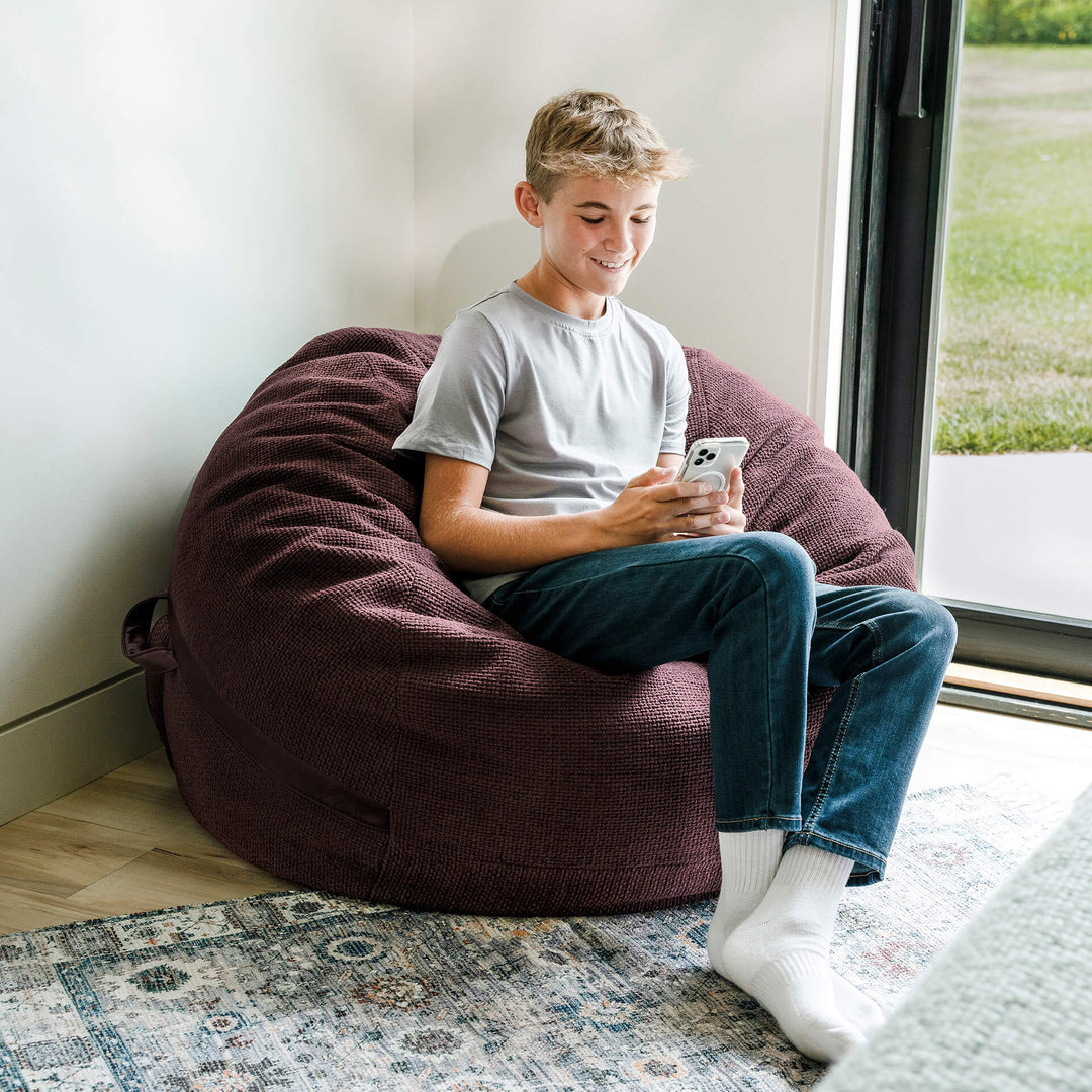 Person sitting on a bean bag chair using a smartphone indoors. #color_merlot-cloudweave