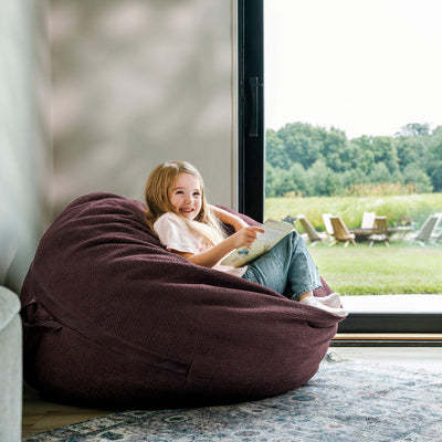 Child sitting in a bean bag chair reading a book by a large window with a view of greenery. 