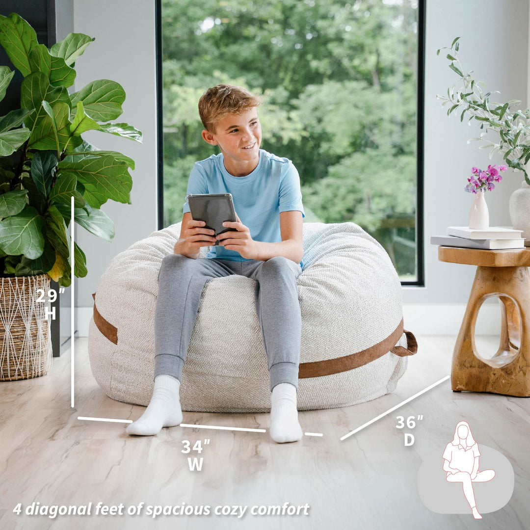 Child sitting on a bean bag chair with dimensions displayed, in a room with plants and a window. #color_sandstone-textured-boucle